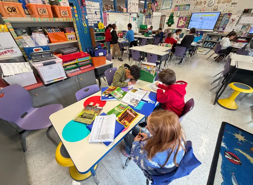 In a classroom, three students read and write by hand at a table. In the background, several other students work and talk.