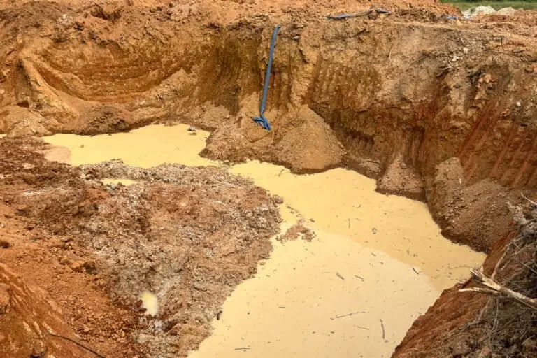 Muddy yellow water against the red earth of a mining pit near Kumasi, Ghana, an abandoned shovel just upright from soil on the far side. Image by Élodie Toto for Mongabay.