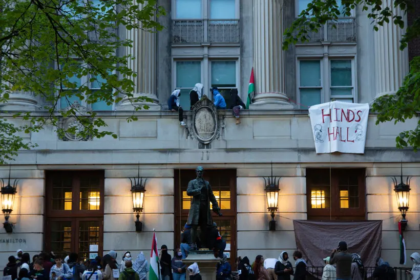 Student protesters gather at Columbia's Hamilton Hall or "Hind's Hall," renamed after Hind Rajab, a six-year-old Palestinian girl who was shot to death with paramedics on an IDF-supplied emergency route. On the right, they hang a banner that says "HIND'S HALL." Protesters sit up high on a facade.