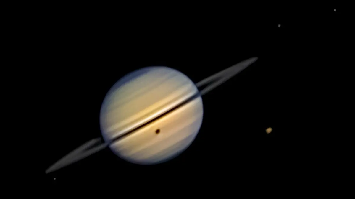 Saturn's largest moon, Titan, casts a small dark shadow over the planet's surface during a rare solar eclipse, while Dione, Tethys, and Enceladus can be seen near the rings. Image: Adam Smadi.