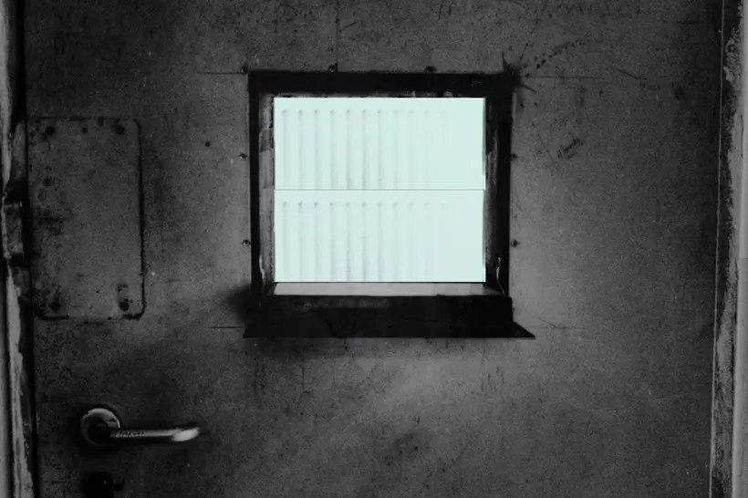 A frightening metal door in black and white shows a small window emanating a harsh bright light.