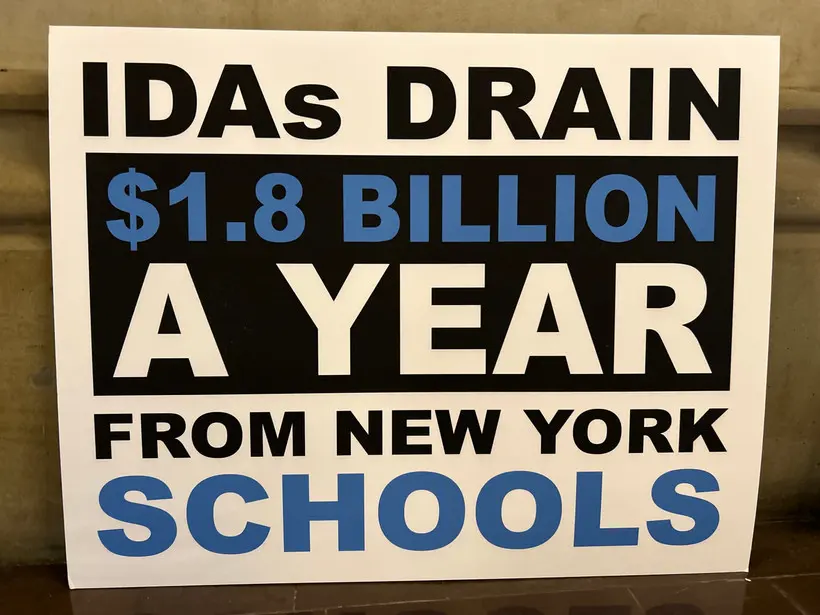 A sign reads "IDAs DRAIN $1.8 BILLION A YEAR FROM NEW YORK SCHOOLS."