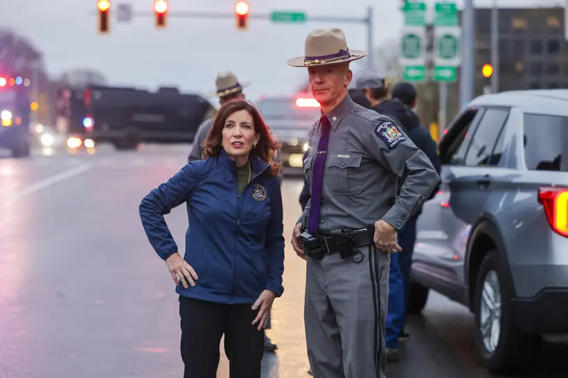 Governor Kathy Hochul stands with a hand on her hip next to a New York state trooper on a cloudy day.