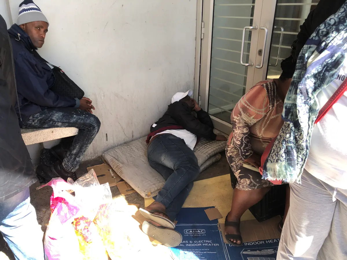 A man at the front of the line asleep on a thin mattress and cardboard sheets.